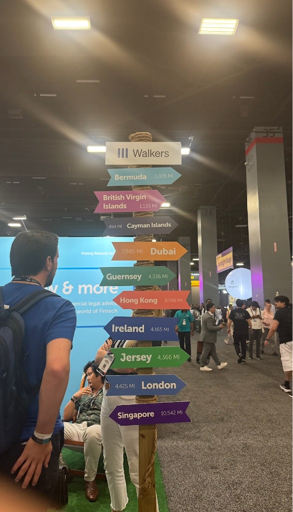 Convention floor signpost with arrows to global financial centers including Cayman Islands 454 miles from Miami